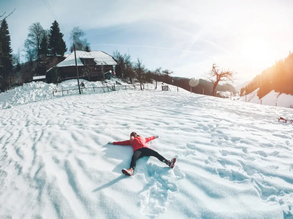 Young woman making a snow angel on a fresh snowy landscape with a cozy mountain house in the background, having fun during a winter vacation in Scandinavia.