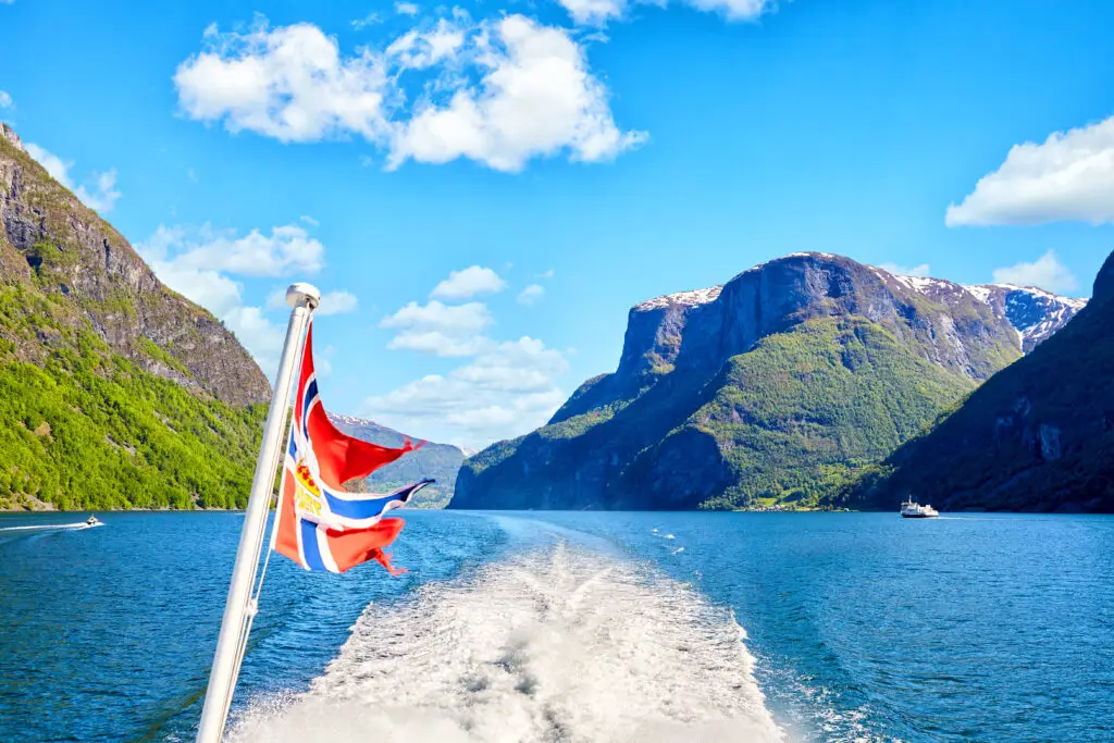 Norwegian flag waving on the stern of a ferry cruising through Sognefjord, Norway, with scenic mountainous landscape in the background.