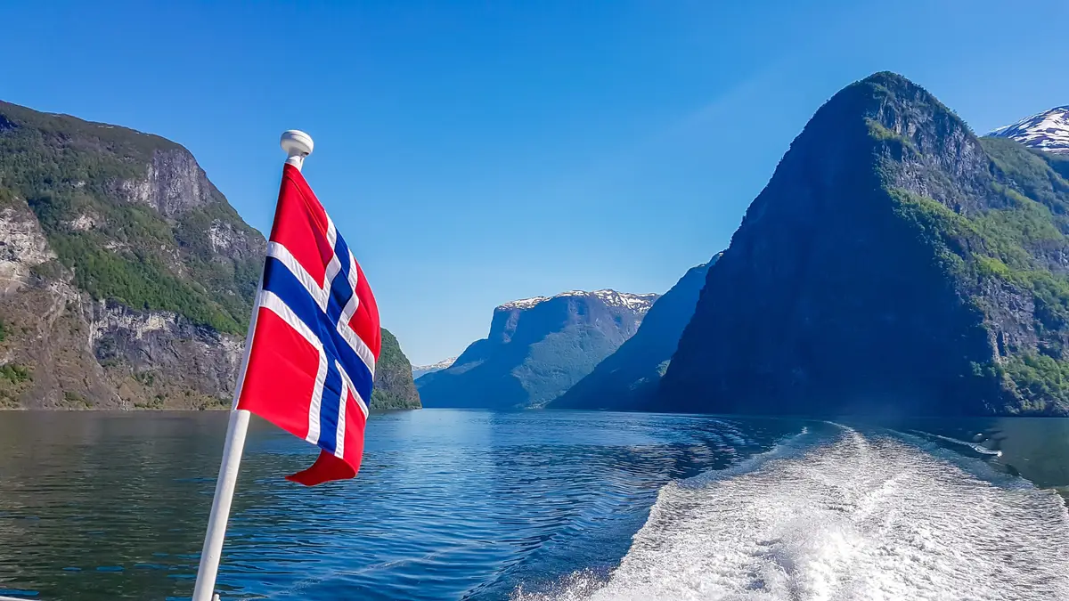 Norwegian flag waving on a ship in a fjord with foamy water trails, surrounded by towering green mountains under a clear blue sky, showcasing Norway's natural beauty.