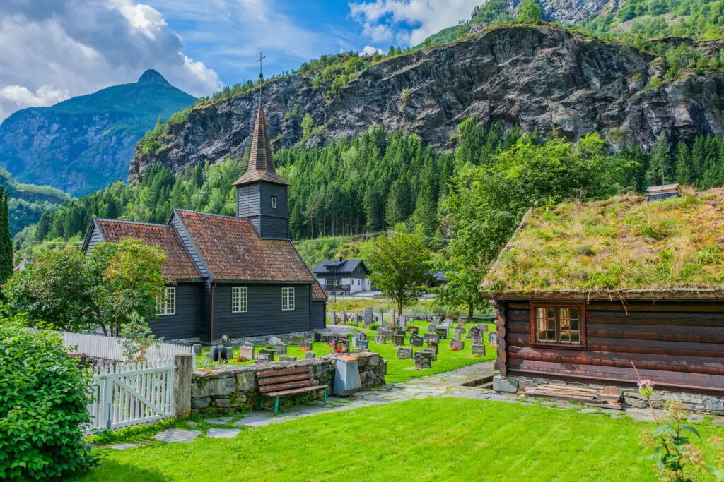 Traditional Norwegian church with grass roof in a scenic village along the fjord journey from Bergen to Flam, surrounded by mountains and lush greenery.