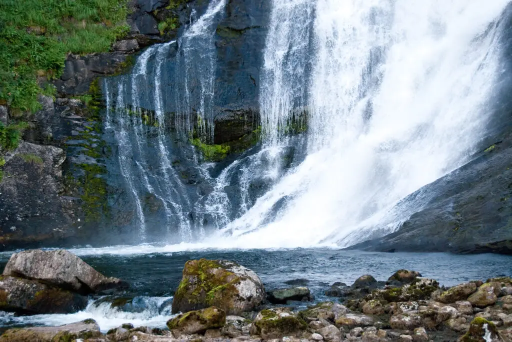 Majestic Huldefossen Waterfall in Norway with lush greenery and cascading waters over dark rocks.