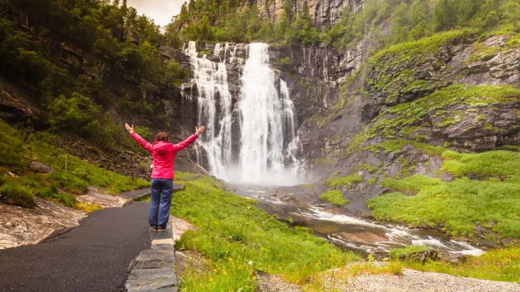 Tourist woman in red jacket raising her arms at Skjervsfossen Waterfall, Norway, showcasing the majestic natural beauty and lush greenery surrounding the cascade.