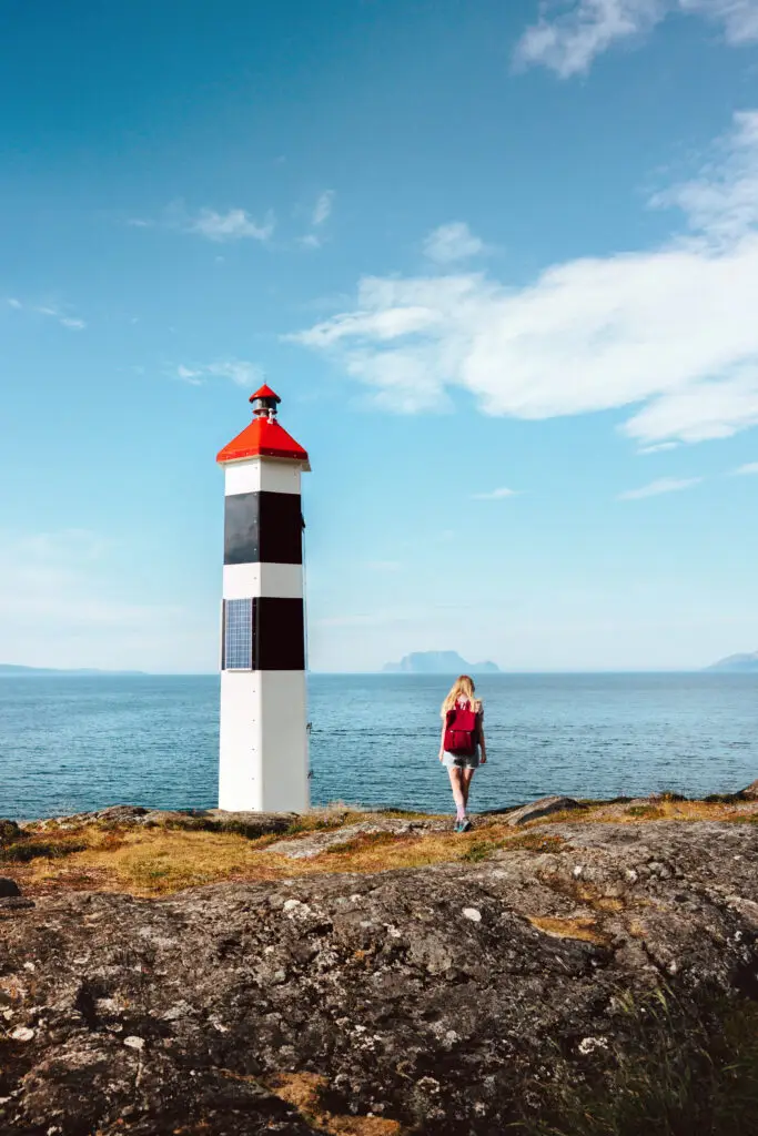 Woman walking towards Lyngstuva lighthouse in Norway, enjoying scenic ocean views during a solo hiking trip, embodying a healthy lifestyle and adventure in summer.