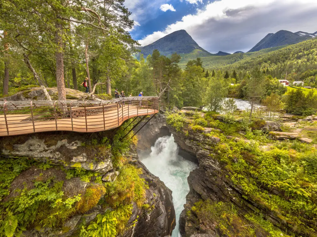 Tourists enjoying the scenic Gudbrandsjuvet waterfall from a viewing platform in Norway, surrounded by lush greenery and mountain landscape.