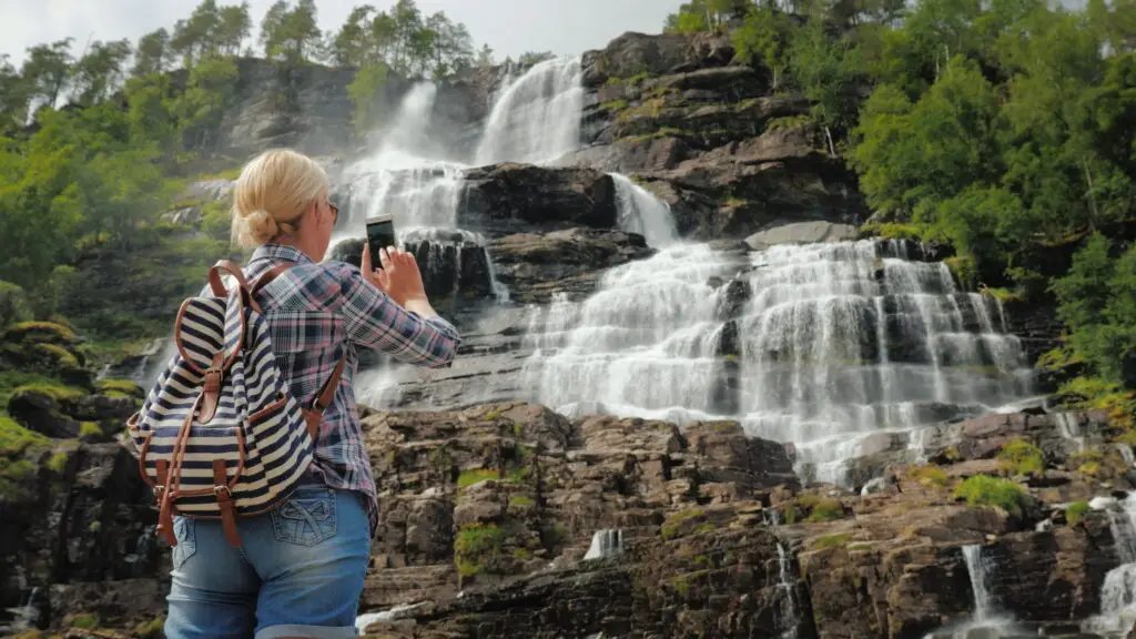 Tourist photographing Norway's highest waterfall, believed to have rejuvenating properties, with a smartphone, showcasing the scenic beauty and natural splendor of Norway's landscapes.