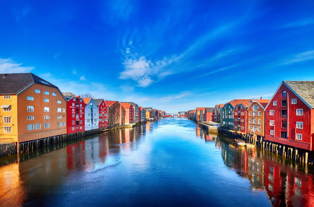Colorful wooden houses on stilts along Nidelva river under a bright blue sky in Trondheim, Norway, reflecting cultural architecture and scenic water views perfect for tourism.