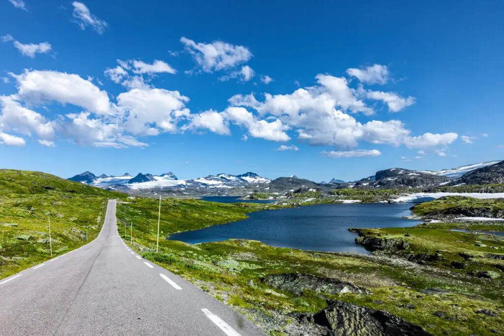 Scenic view of Route 55 across Sognefjell in Jotunheimen, Norway, showcasing a clear road leading through lush landscapes with snow-capped peaks and vibrant blue lakes under a sunny sky.