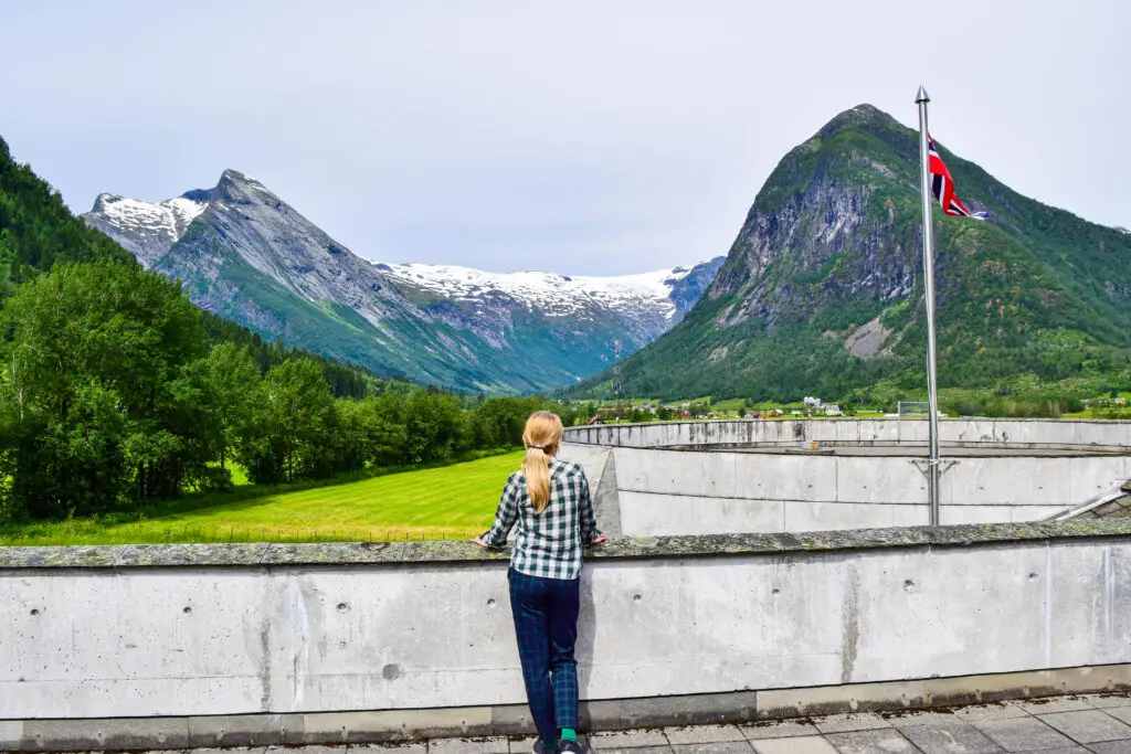 Young woman admiring the breathtaking mountain landscape and glaciers from the Glacier Museum in Fjaerland, Norway, with the Norwegian flag fluttering in the foreground.