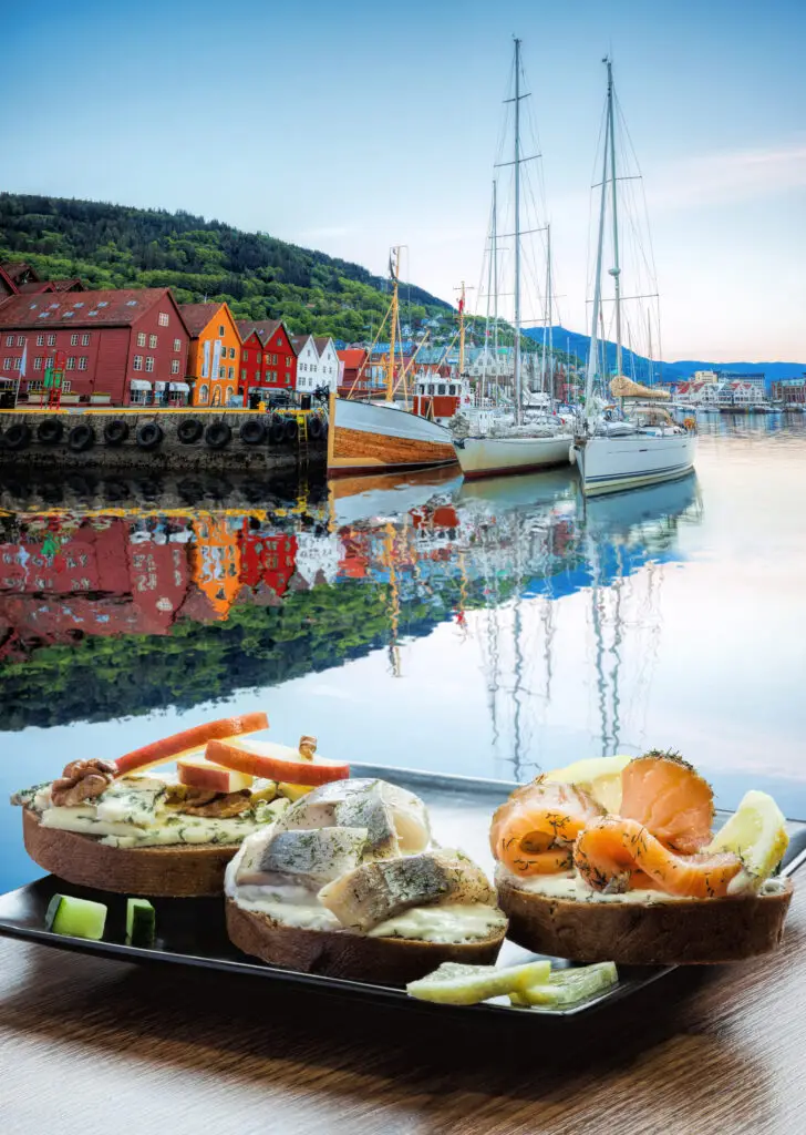 Assorted Scandinavian sandwiches on a wooden table with a scenic view of colorful Bryggen street and sailboats reflected in the calm waters of Bergen, Norway, a UNESCO World Heritage Site.