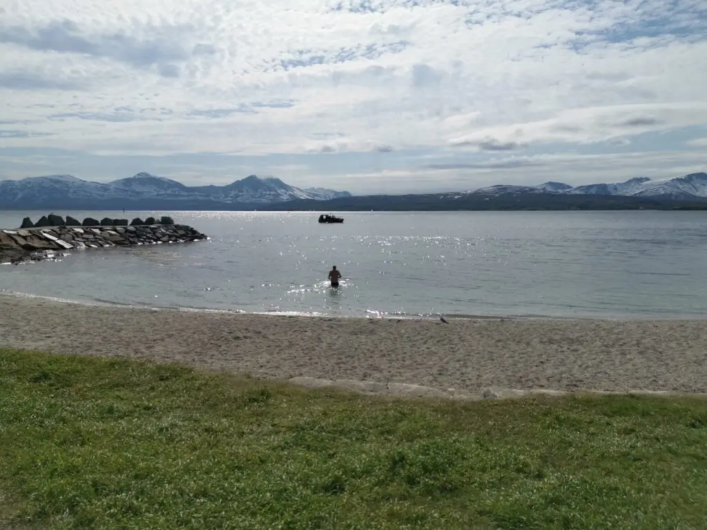 Summer view of Telegrafbukta beach in Tromsø, Northern Norway, featuring a serene seascape with snowy mountains in the background and a person wading in the sparkling water.
