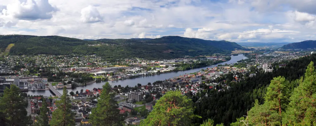 Panoramic view from Spiralen lookout capturing the scenic city of Drammen by the Drammenselva River in Norway, showcasing lush greenery, urban landscape, and a vivid blue sky with clouds.