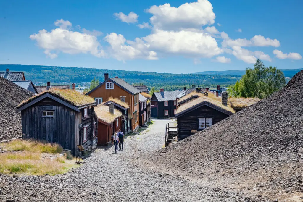 Tourists walking down a cobblestone street in Røros, Norway, surrounded by historical wooden buildings with grass roofs, highlighting the city's heritage as a former copper mining hub.