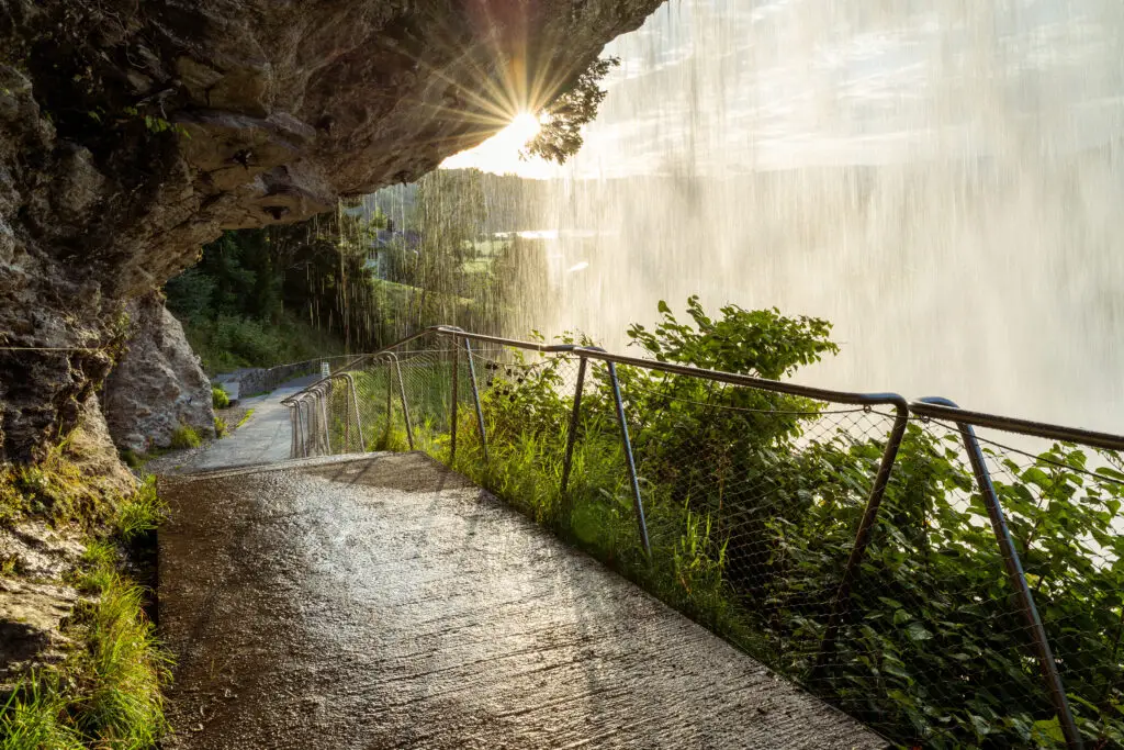 Pathway under the cascading Steinsdalsfossen waterfall in Norheimsund, Norway, showcasing a sunbeam piercing through water spray.
