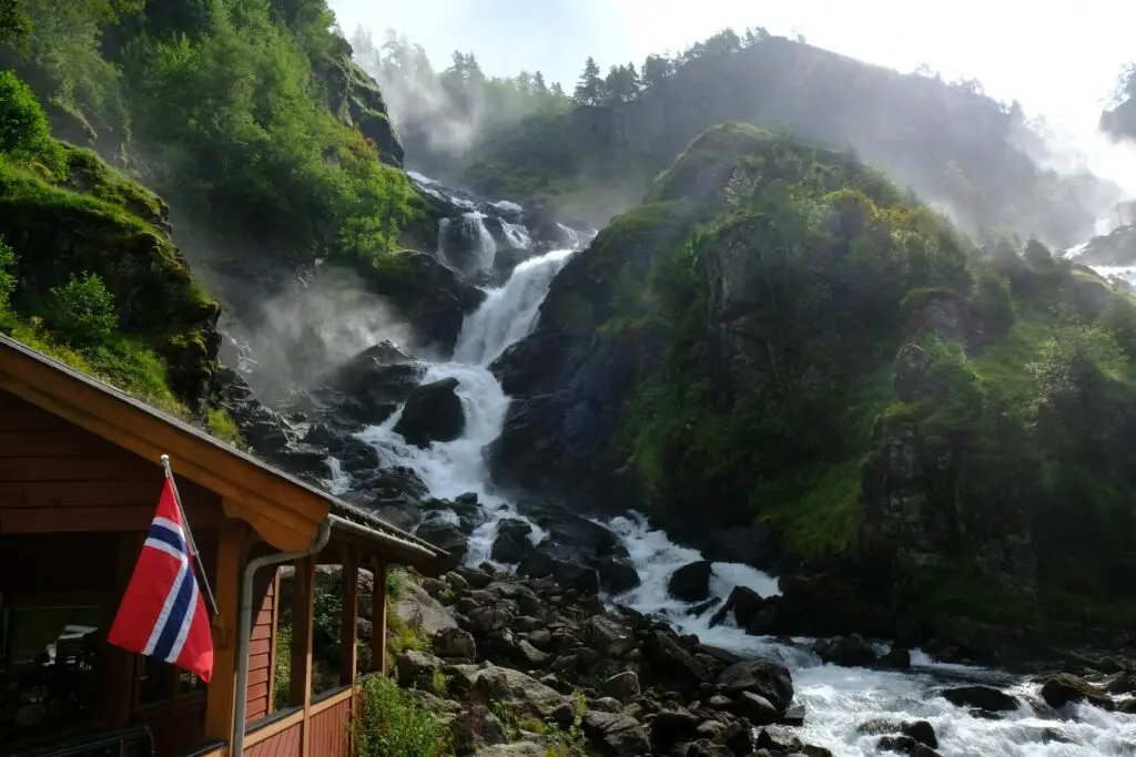 Scenic view of Latefossen and Espelandsfossen waterfalls in Norway with mist rising around lush greenery, featuring a Norwegian flag on a traditional wooden building.