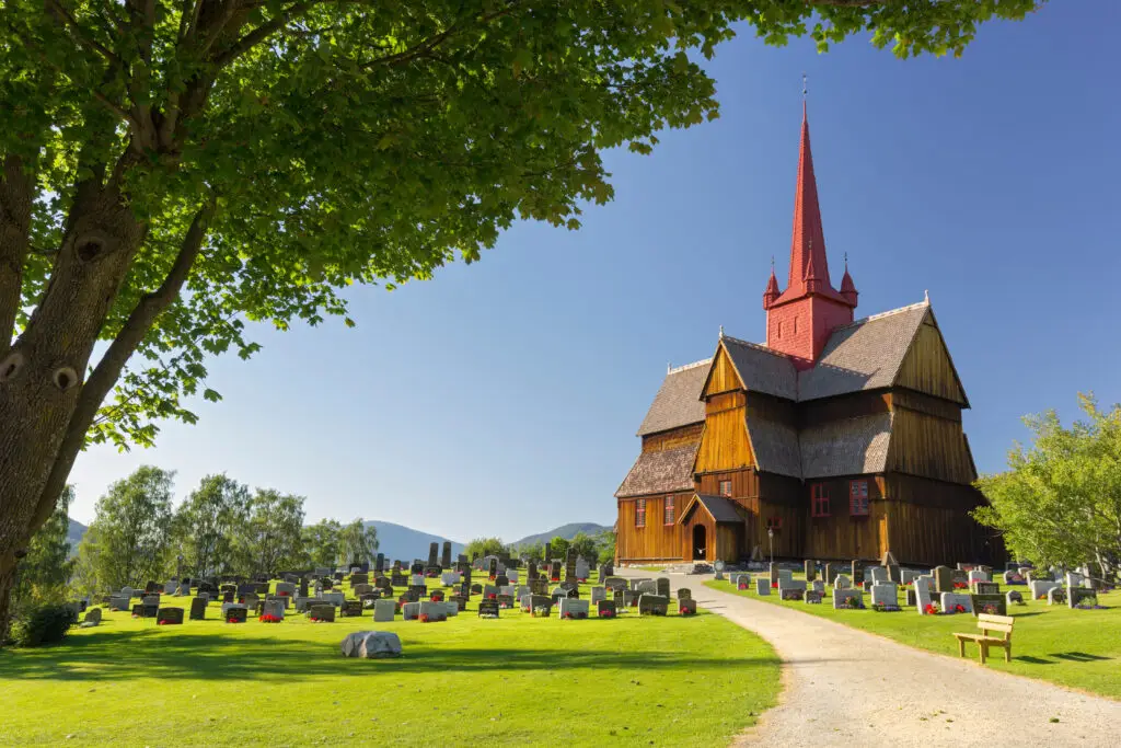 Stabkirche von Ringebu, an ancient wooden stave church in Gudbrandsdalen, Oppland, Norway, surrounded by a cemetery with colorful flowers on graves and lush green trees under a clear blue sky.