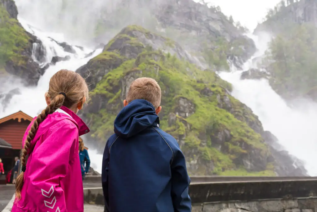 Kids gazing at majestic waterfall in Norway, surrounded by lush greenery and mist.