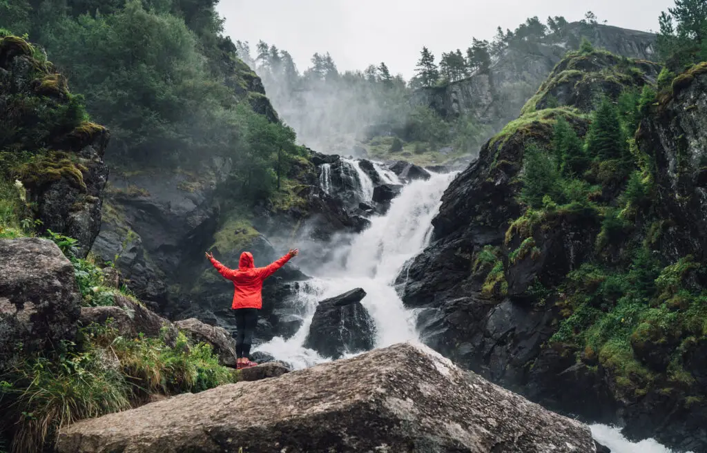 Woman in red jacket celebrating the majestic Låtefossen Waterfall in Norway amidst misty rocky landscape